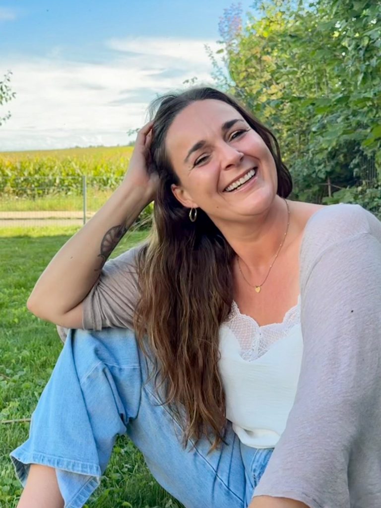 Junge Frau sitzt lächelnd im Grünen, umgeben von Bäumen und einem klaren Himmel.